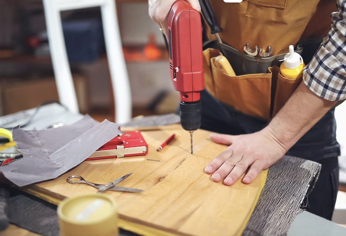 Handwerker mit Werkzeuggürtel bohrt an einem Holzbrett, Werkzeug liegt griffbereit.